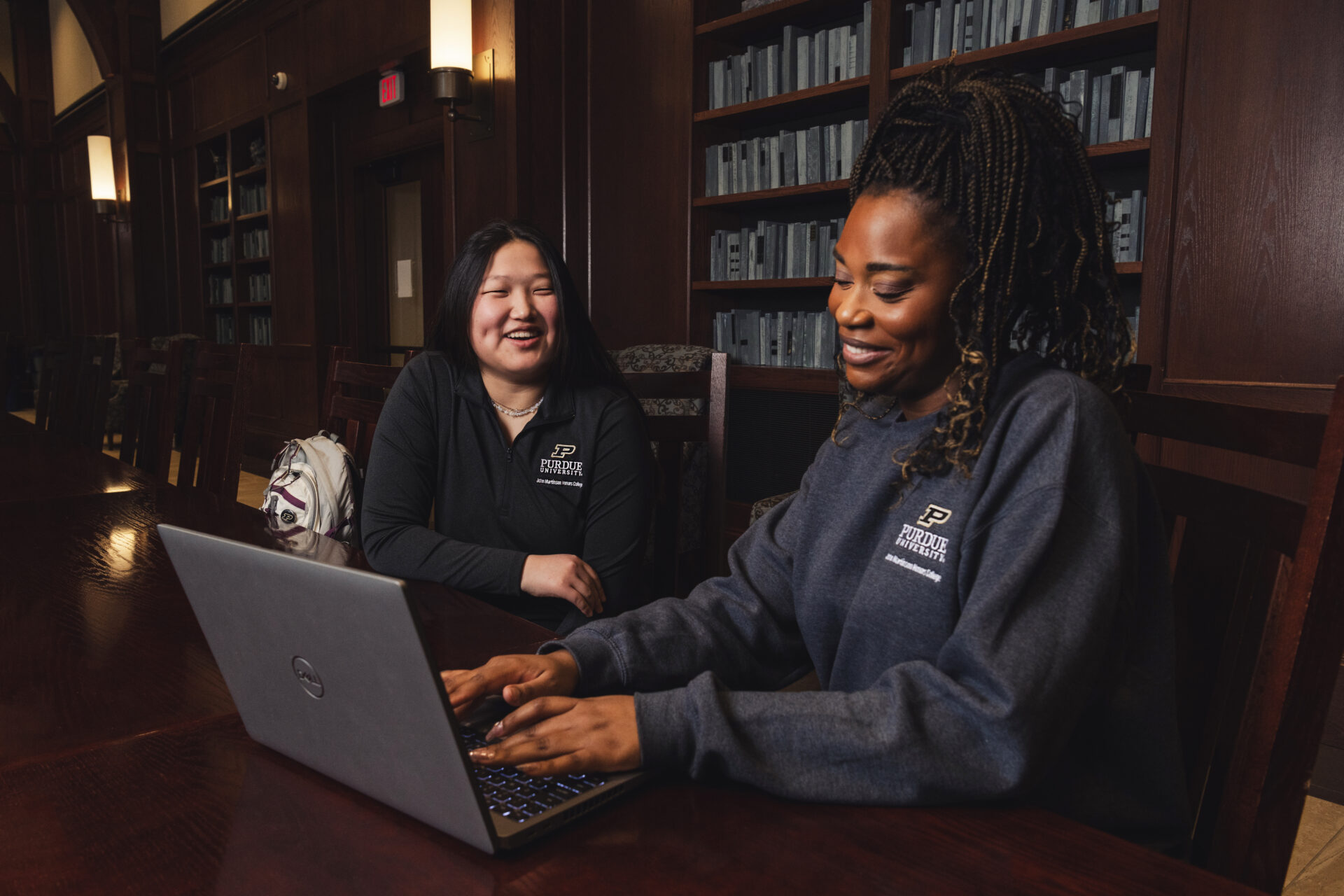 Two females sitting next to each other in a library working on a laptop.