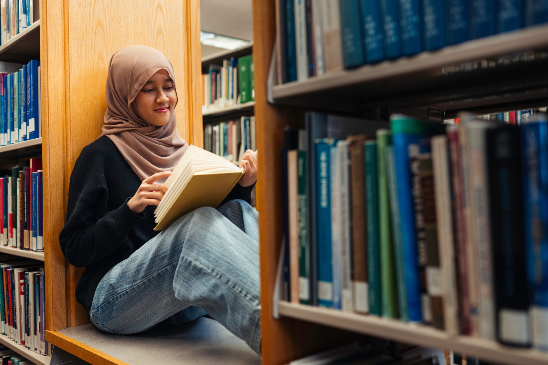 Female sitting between two library shelves reading a book.