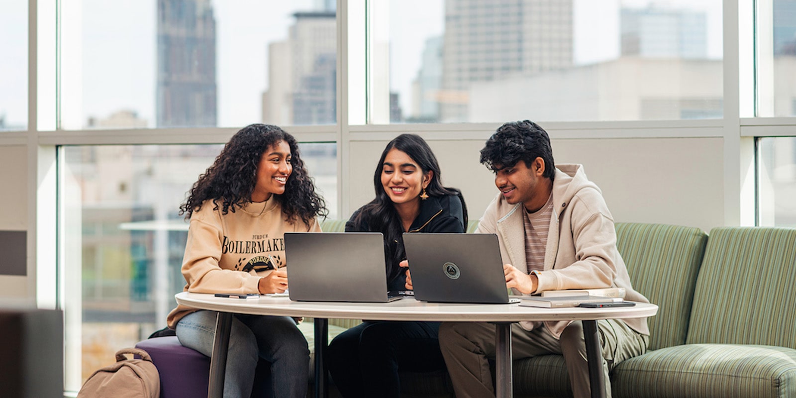 Three smiling students sit at a table looking at laptops in front of a large window with a city view.