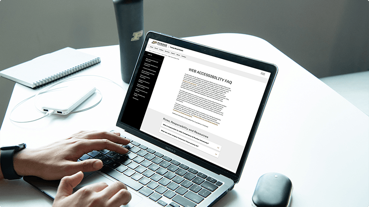 A person using a laptop that displays the Purdue Brand Studio Web Accessibility FAQ page, with a Purdue water bottle and notebook on the desk.