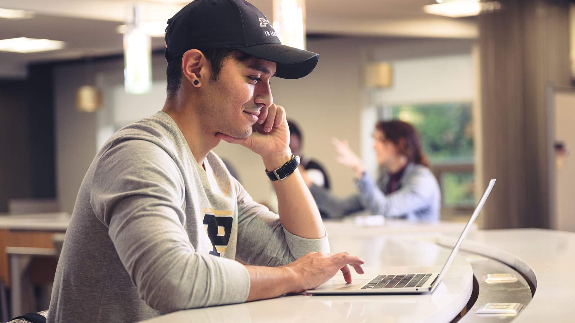 A student wearing a Purdue baseball cap smiles while working on a laptop at a counter.