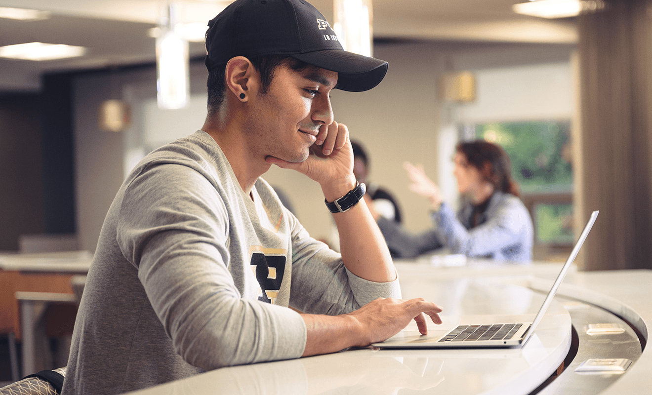 A student wearing a Purdue baseball cap smiles while working on a laptop at a counter.