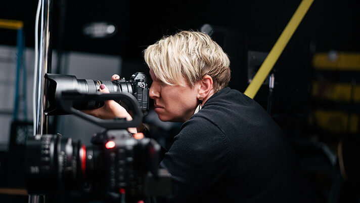 A photographer with short blonde hair looks through a camera viewfinder in a dark studio setting.