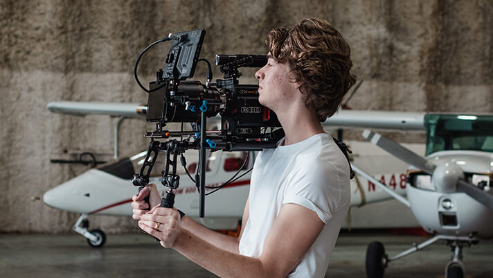 A videographer operating a shoulder-mounted camera rig inside an airplane hangar with small aircraft in the background.