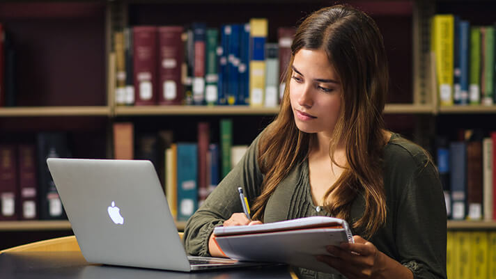 A student writing in a notebook while looking at a laptop in a library.