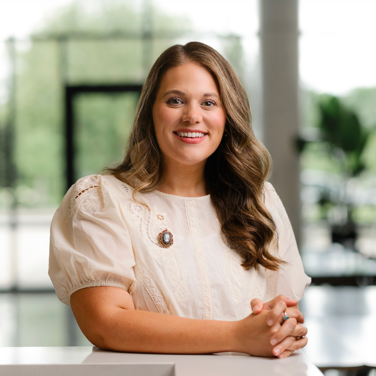 A person with long, wavy hair smiles warmly while seated at a white table in a bright, modern space with large windows.
