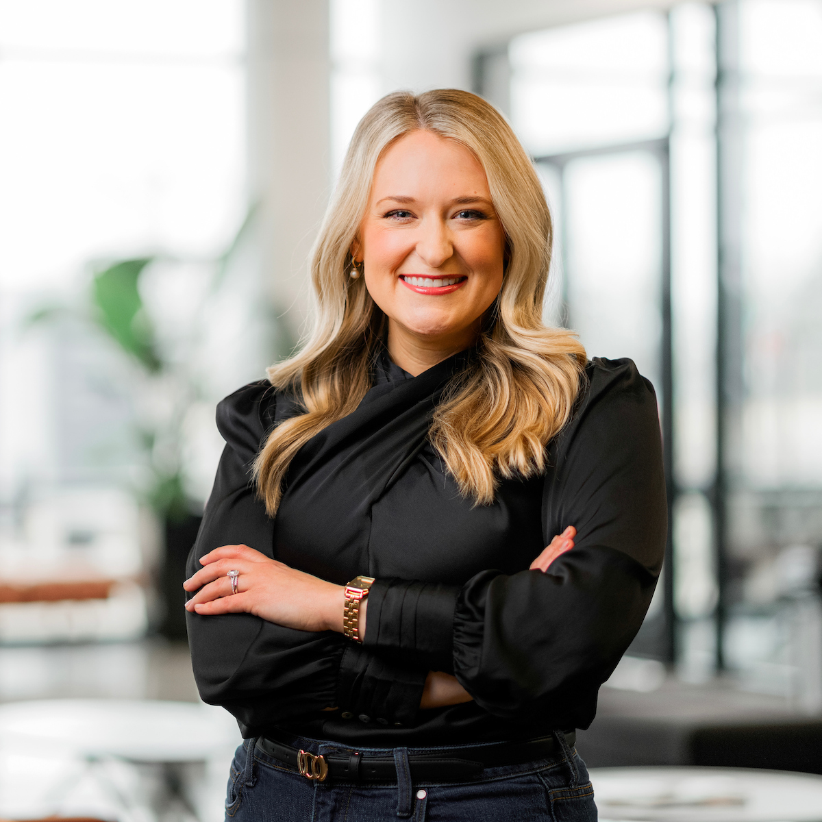 A professionally dressed individual with blonde hair stands confidently with arms crossed in a modern, well-lit office space.