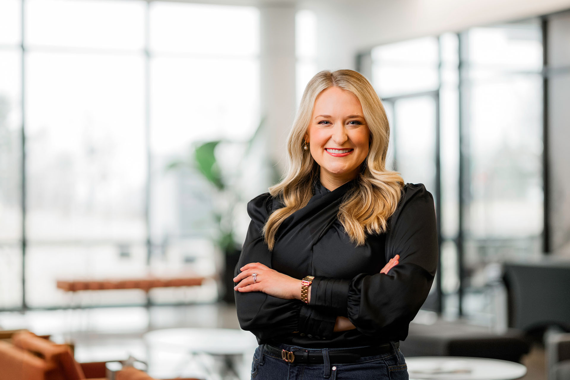 A professionally dressed individual with blonde hair stands confidently with arms crossed in a modern, well-lit office space.