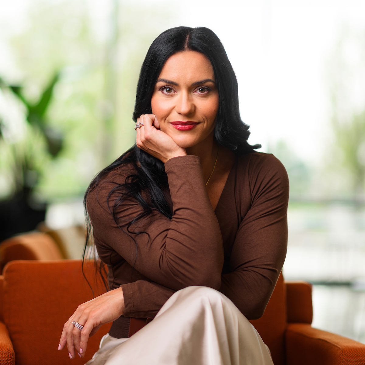 A person with long dark hair sits thoughtfully on an orange couch in a bright, modern space with large windows and blurred greenery outside.