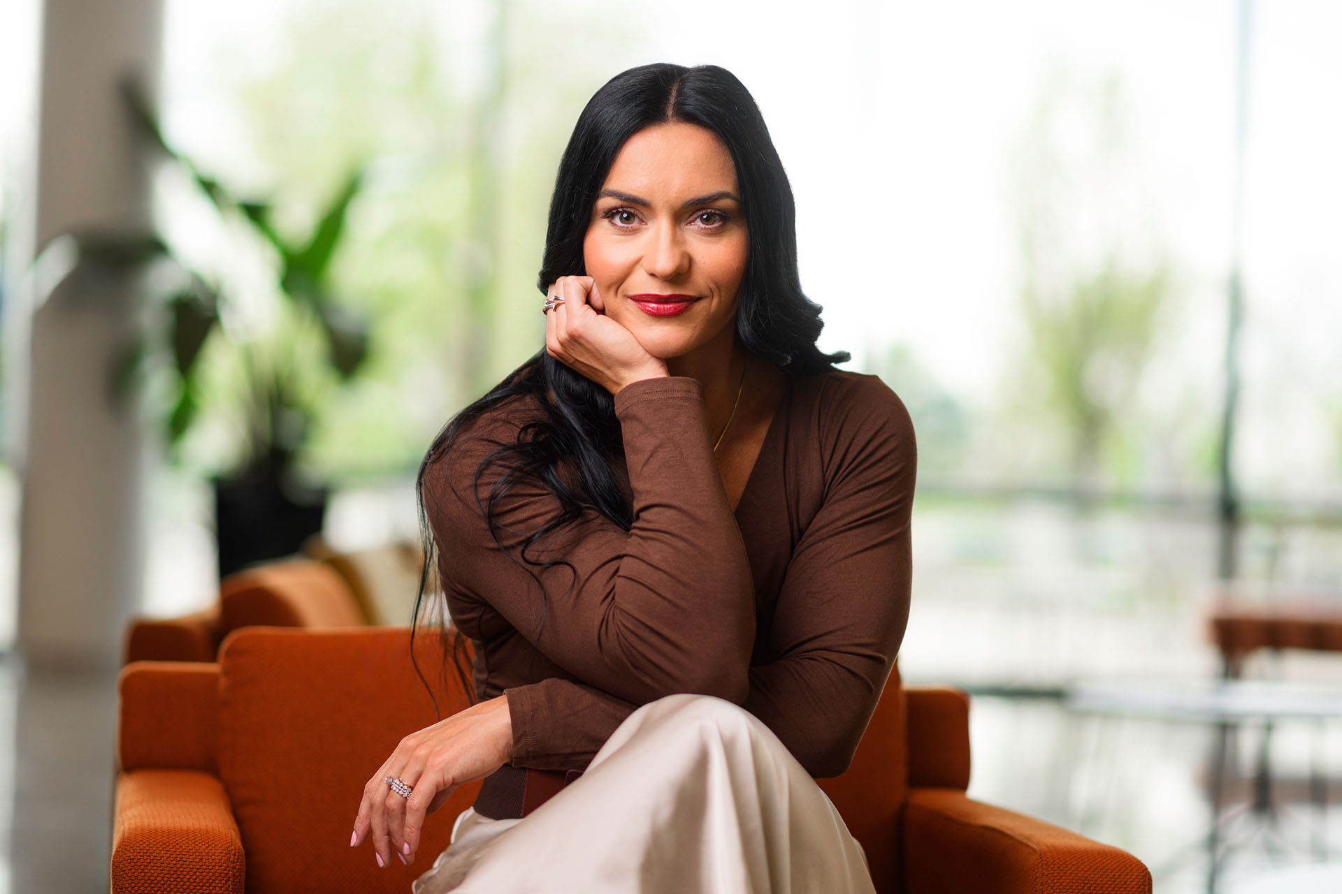 A person with long dark hair sits thoughtfully on an orange couch in a bright, modern space with large windows and blurred greenery outside.