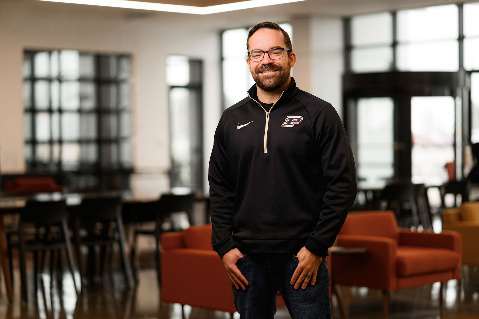 A person wearing a black athletic jacket with a university logo stands smiling in a modern, well-lit indoor space with large windows and orange seating.