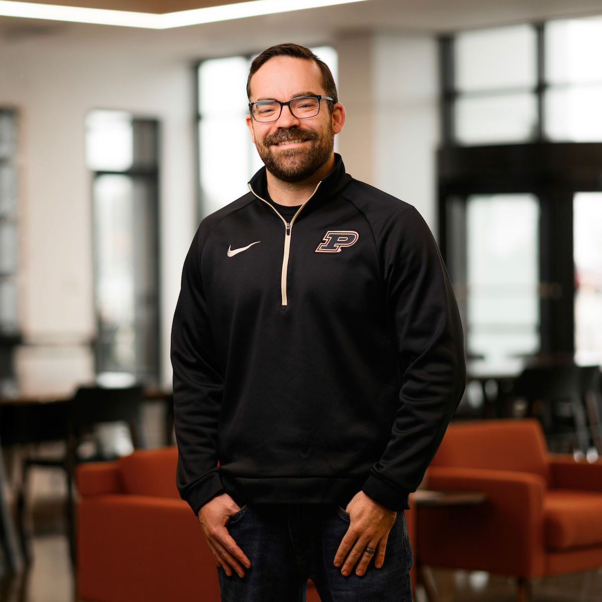 A person wearing glasses and a black athletic jacket with a logo stands smiling in a modern, well-lit space with large windows and furniture.