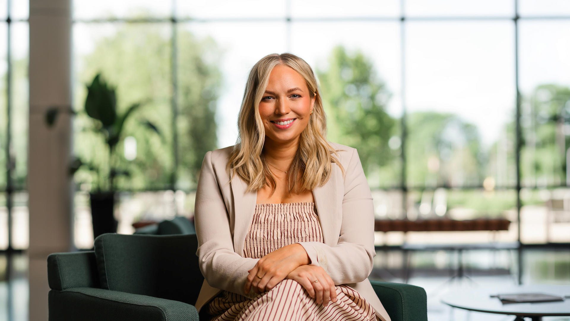 A smiling person with blonde hair sits in a modern chair, wearing a light-colored blazer and striped dress, with large windows and greenery visible in the background.