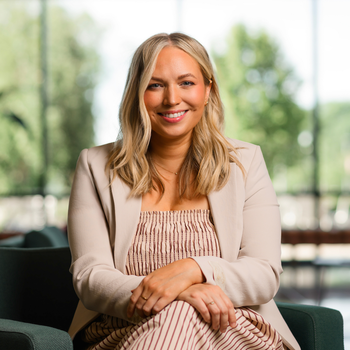A smiling person with blonde hair sits in a chair, wearing a beige blazer and striped dress, with a blurred green outdoor background.