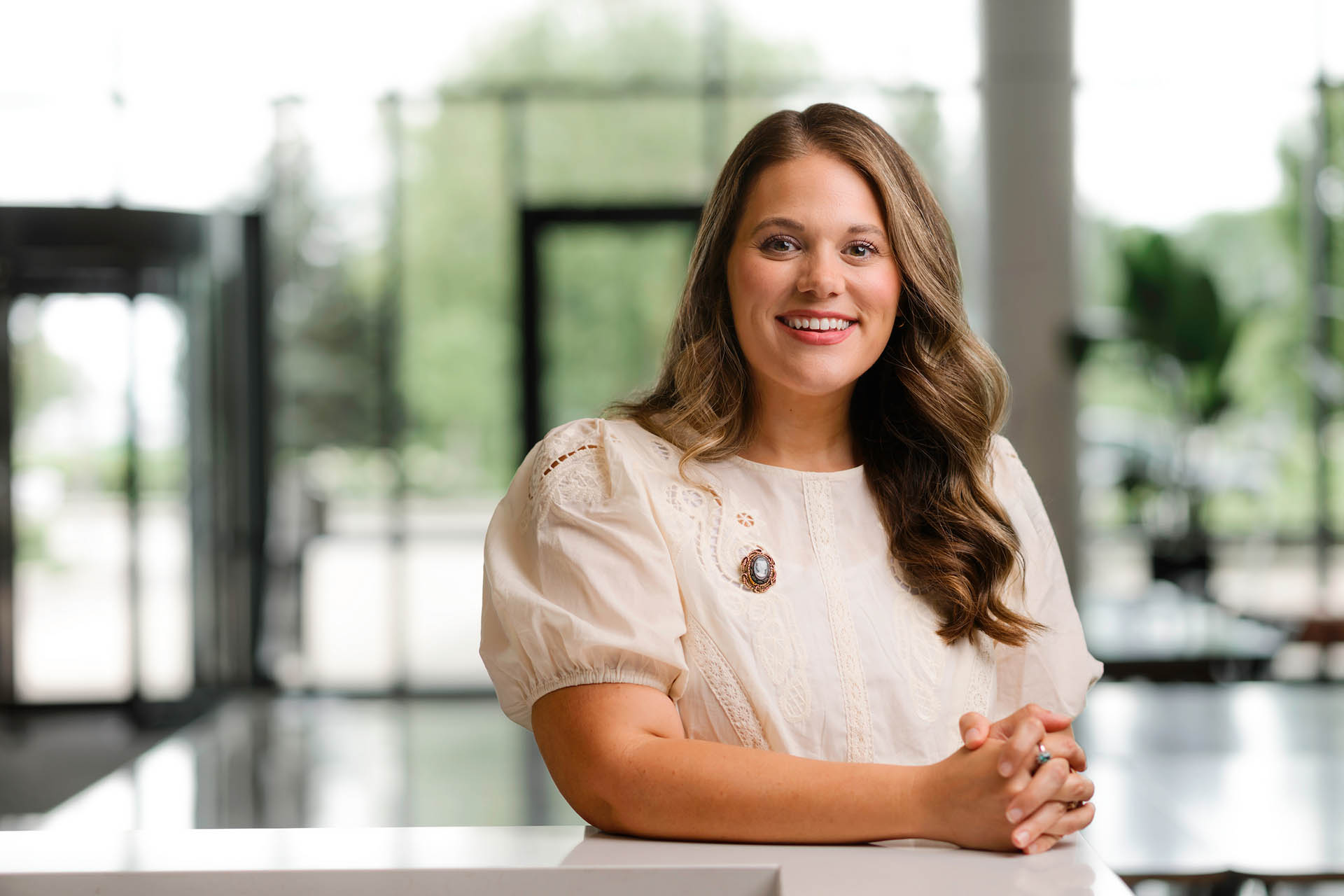 A person with long, wavy hair smiles warmly while seated at a white table in a bright, modern space with large windows.