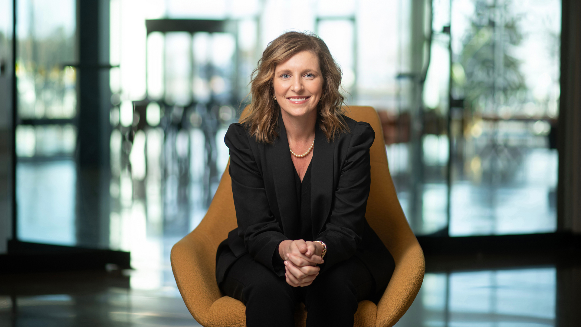 A professionally dressed individual sits in a modern orange chair, smiling at the camera in a brightly lit, contemporary office space.