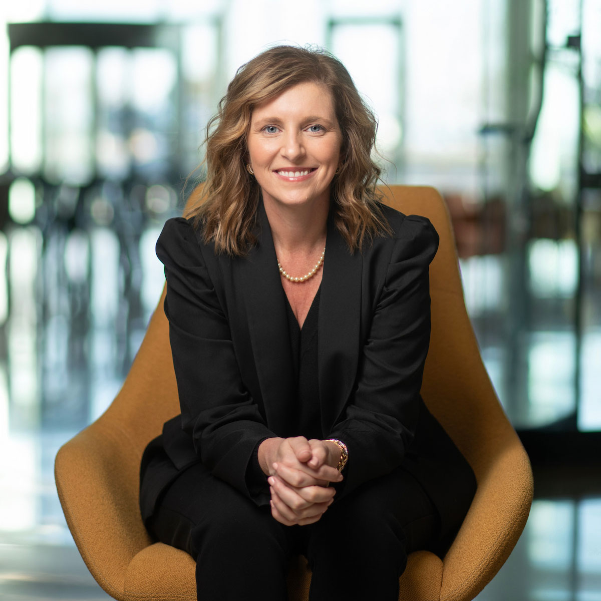 A professionally dressed individual sits in a modern orange chair, smiling warmly at the camera against a softly blurred background.