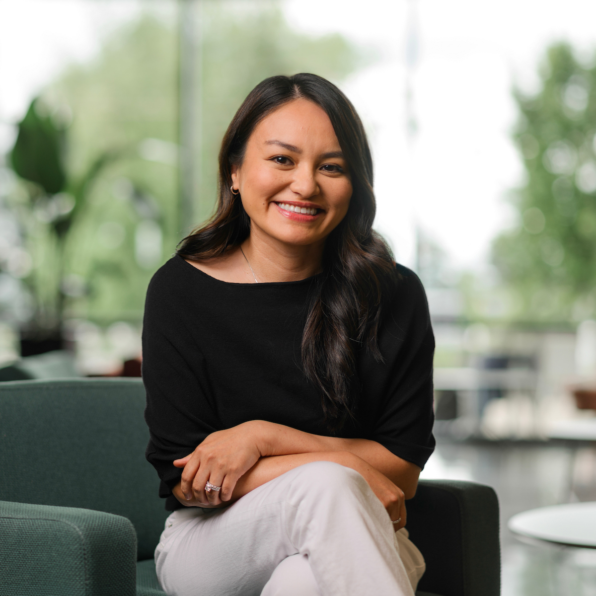 A person with long dark hair sits smiling in a green armchair in a bright, modern space with large windows and blurred greenery outside.