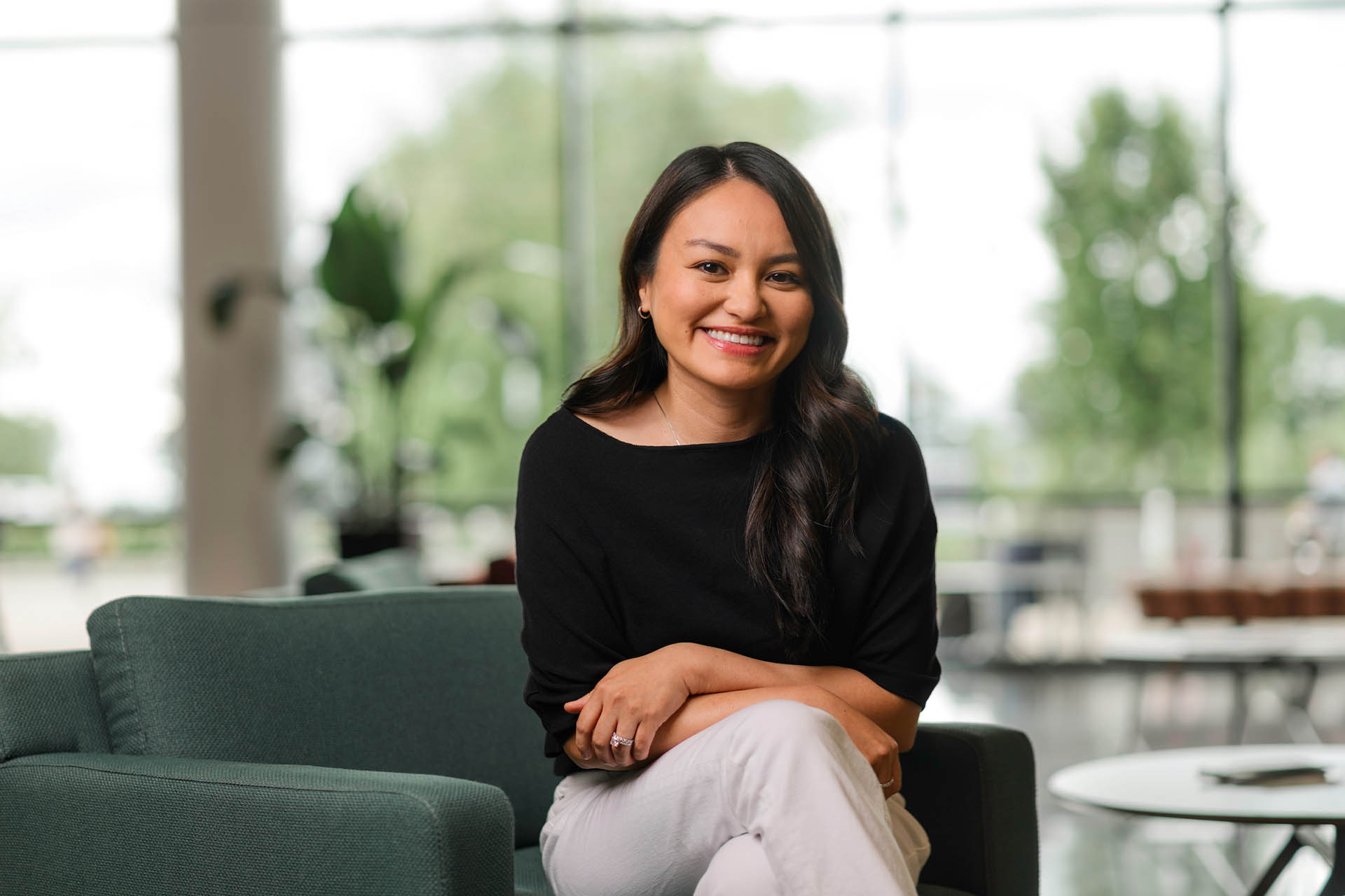 A person with long dark hair sits smiling in a green armchair in a bright, modern space with large windows and blurred greenery outside.