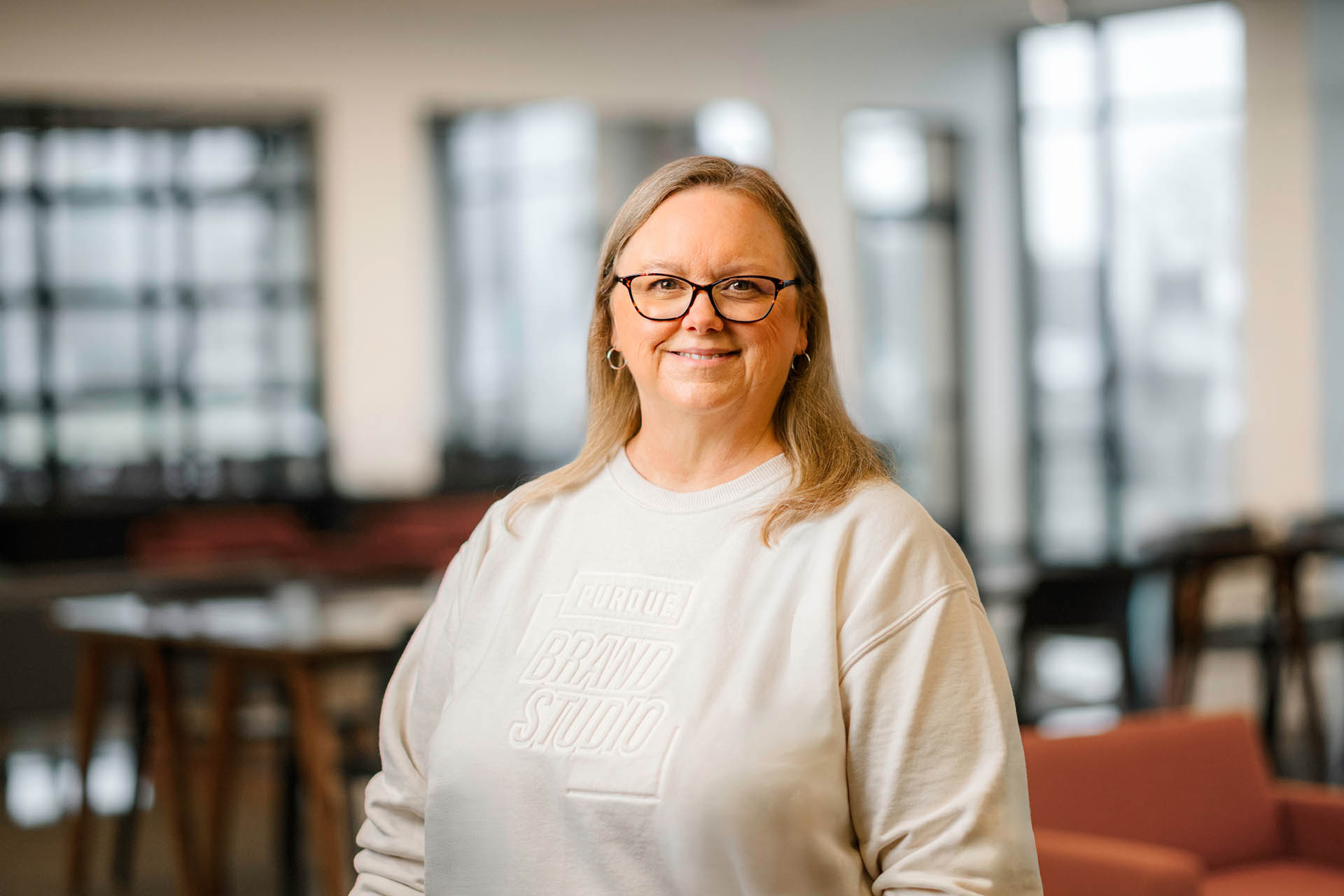 A person with long hair and glasses smiles while wearing a light-colored sweatshirt with "PURDUE BRAND STUDIO" printed on it, standing in a modern, well-lit office space.