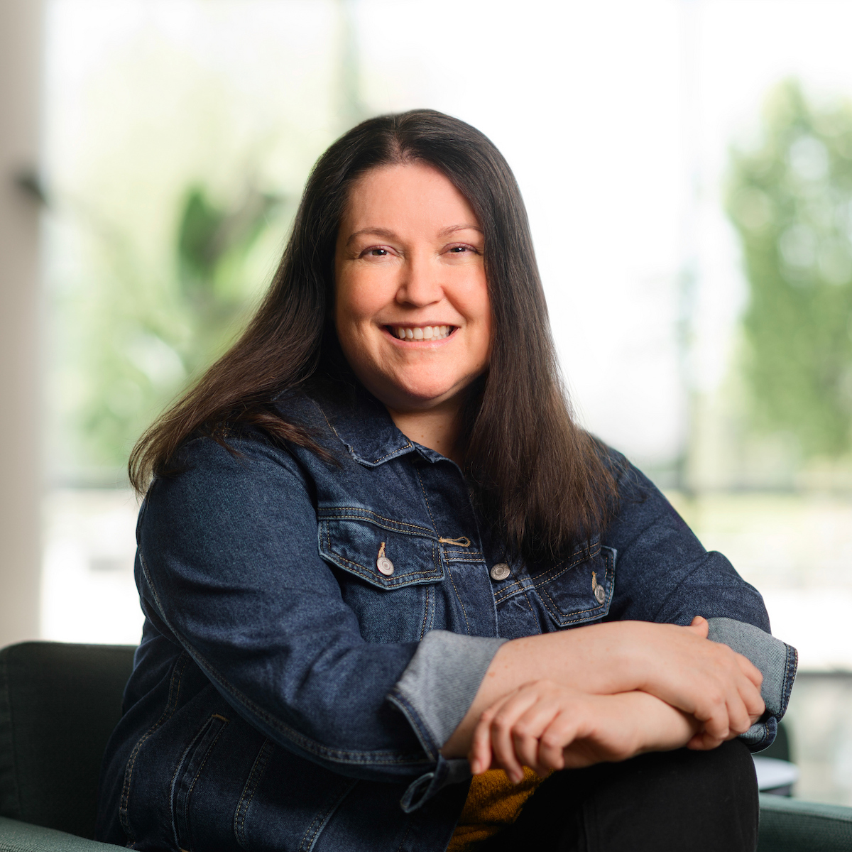 A person with long dark hair smiles warmly while seated, wearing a denim jacket with arms crossed, against a softly blurred background of greenery and light.