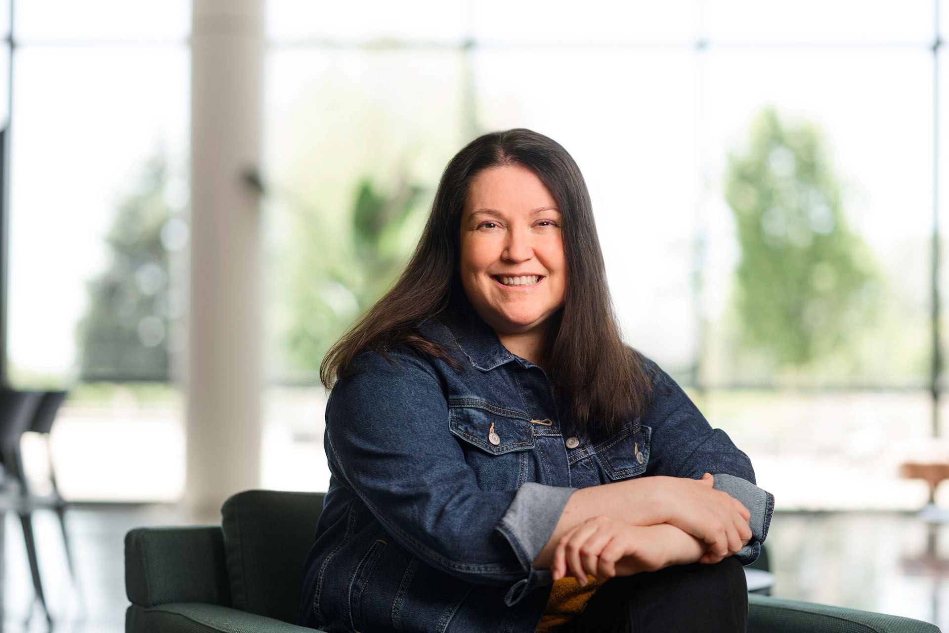 A person with long dark hair, wearing a denim jacket, sits smiling in a modern, brightly lit space with large windows and blurred greenery in the background.