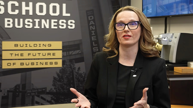 A professional woman with glasses speaking in front of a Purdue School of Business banner.