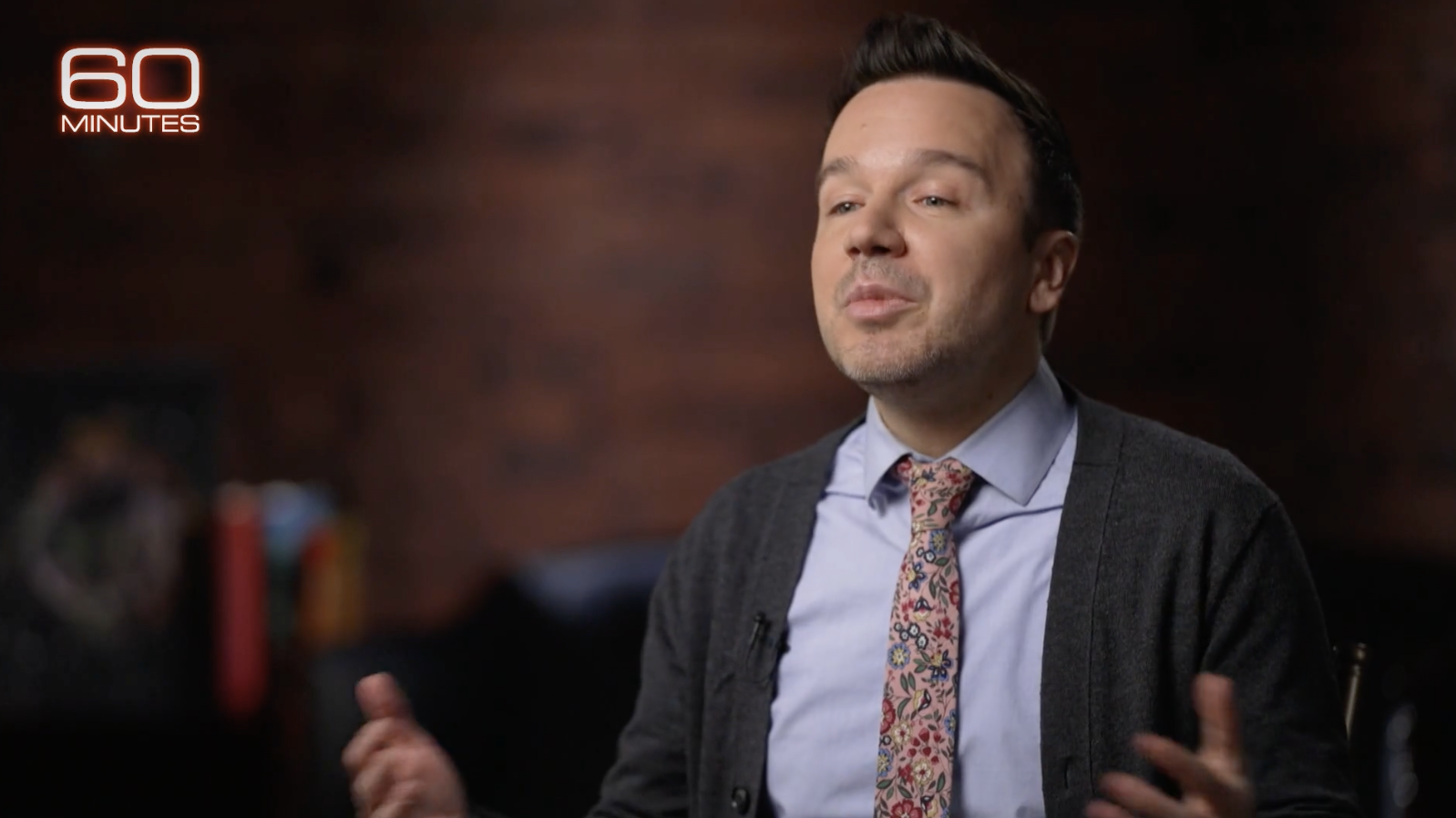 A man wearing a floral tie and cardigan speaking during a 60 Minutes interview.