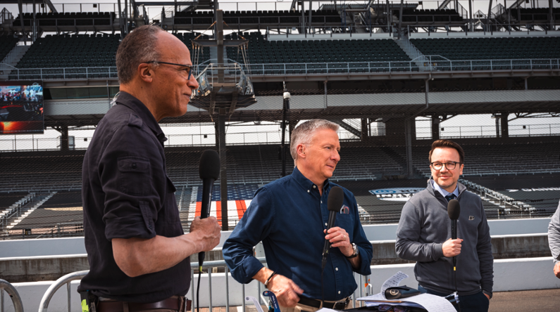 Three men hold microphones at a racetrack stadium.