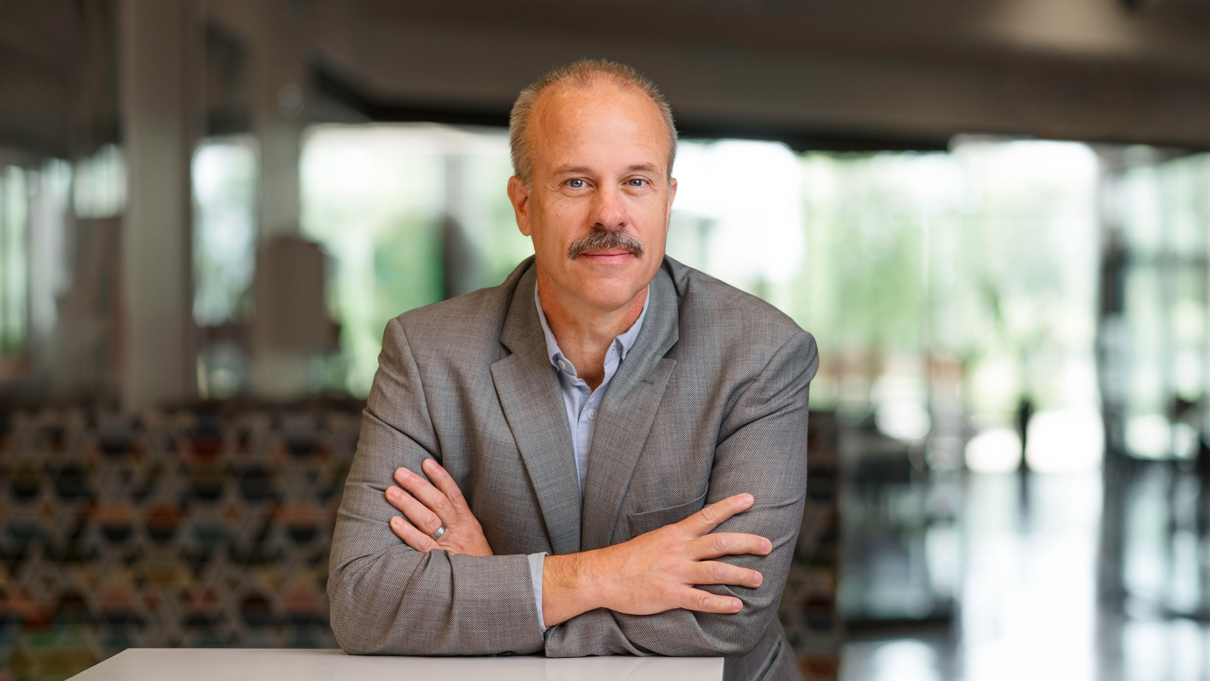A person with a mustache, wearing a gray blazer and light blue shirt, sits at a table with arms crossed, looking directly at the camera in a modern, well-lit indoor space.