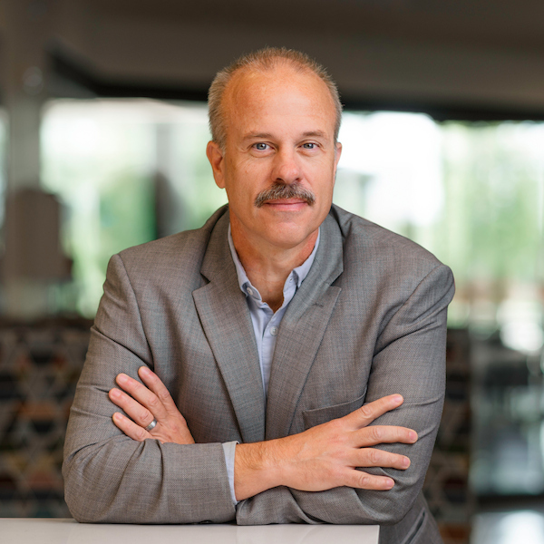 A person with a mustache, wearing a gray blazer over a light blue shirt, sits with arms crossed at a table in a softly lit indoor setting.