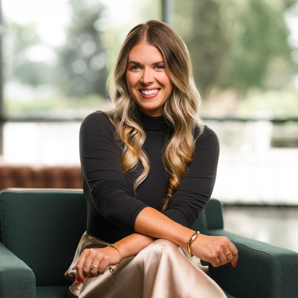 A smiling person with long wavy hair sits on a green armchair, wearing a black turtleneck and a light-colored skirt, with hands clasped and adorned with jewelry.