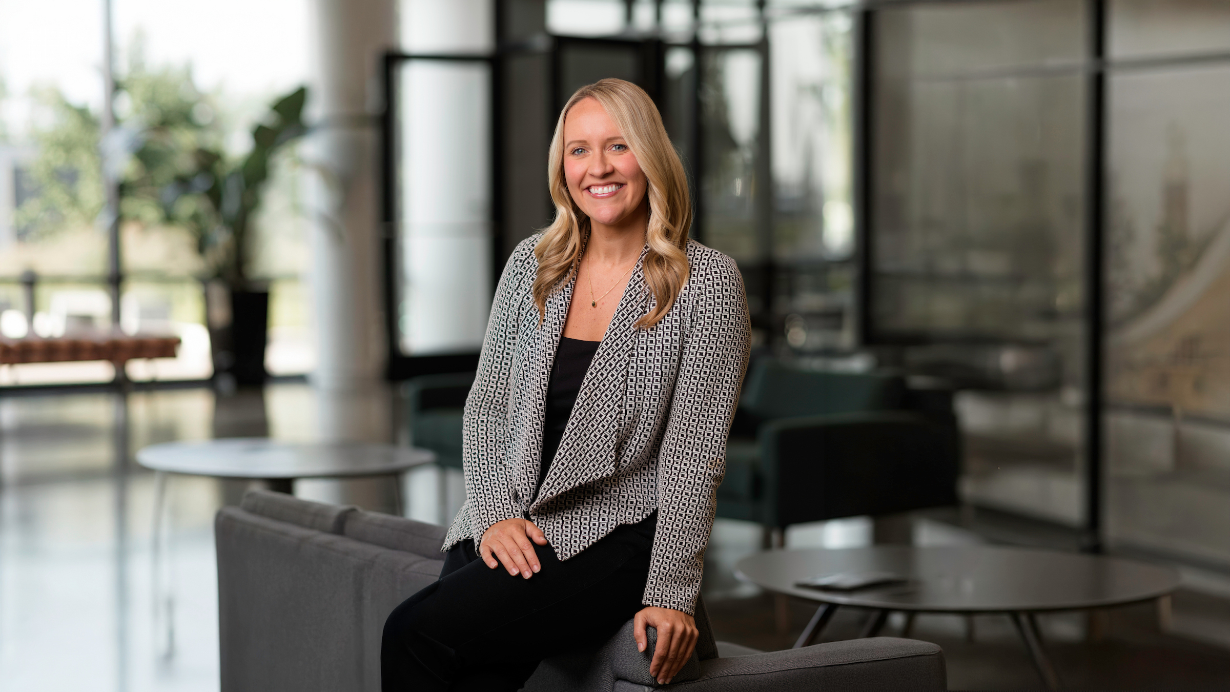 A professionally dressed individual with blonde hair sits smiling on a gray couch in a modern, well-lit office space.