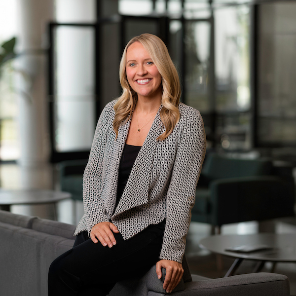 A professionally dressed individual with blonde hair smiles while seated on a gray couch in a modern, well-lit office space.