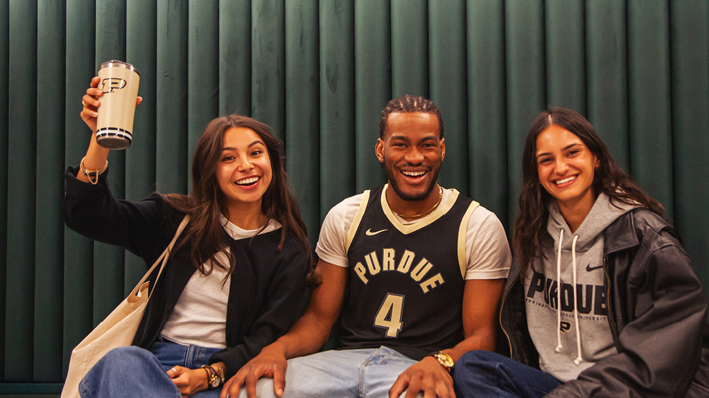 Three smiling Purdue students sitting together against a green wall, with one holding up a Purdue travel tumbler.