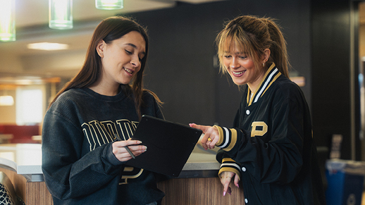 Two students looking at a tablet together indoors while wearing Purdue apparel.