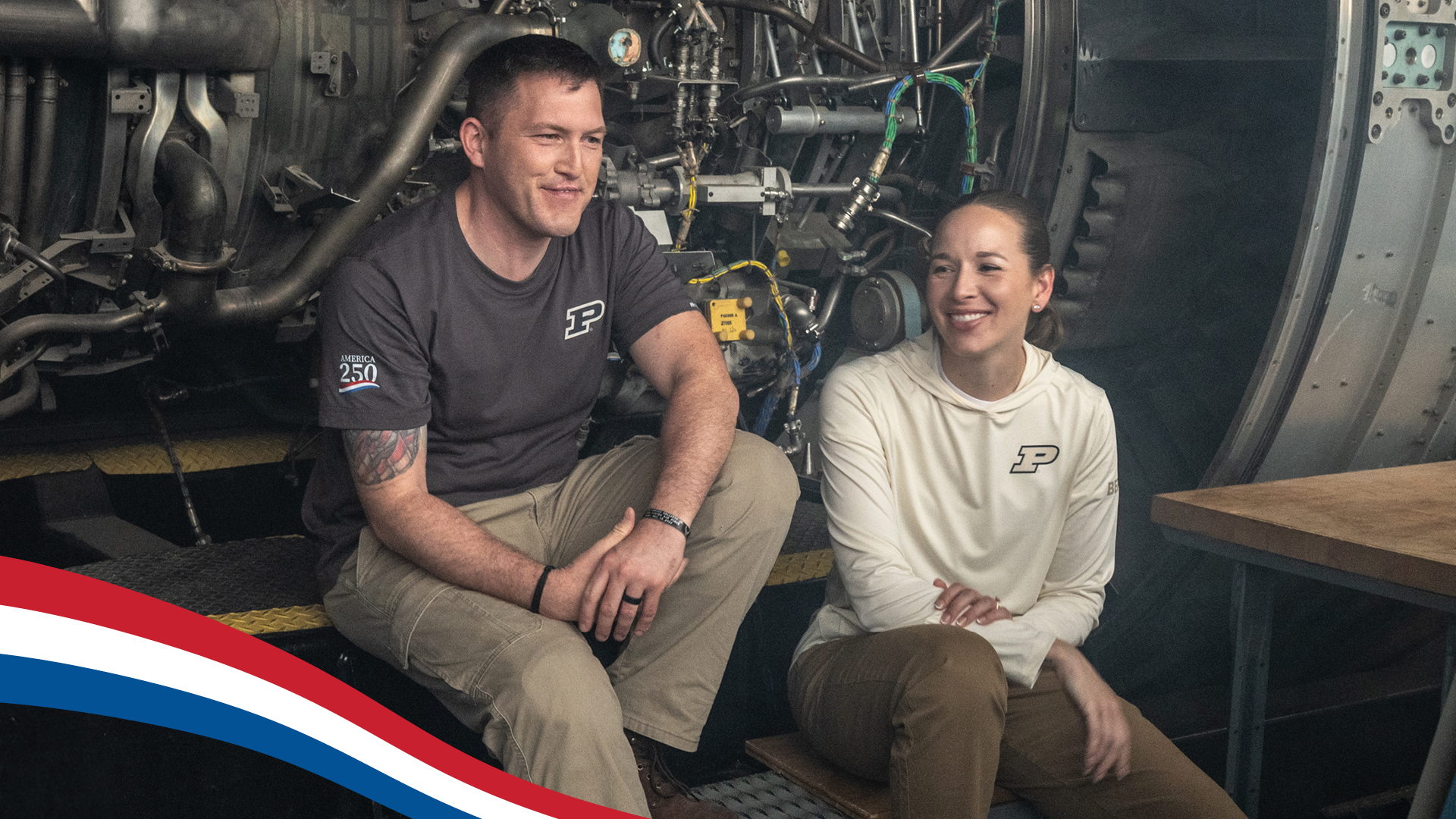 Smiling man and woman wearing Purdue shirts sit in front of a large engine