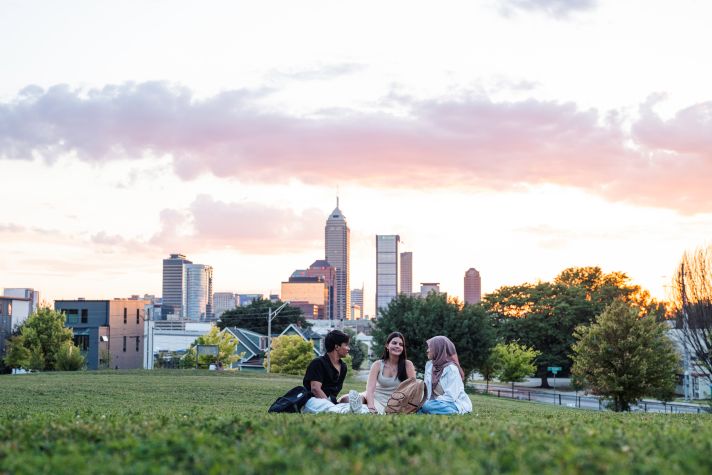 Three people sitting on a grassy area during sunset, chatting with the Indianapolis skyline and colorful clouds in the background.