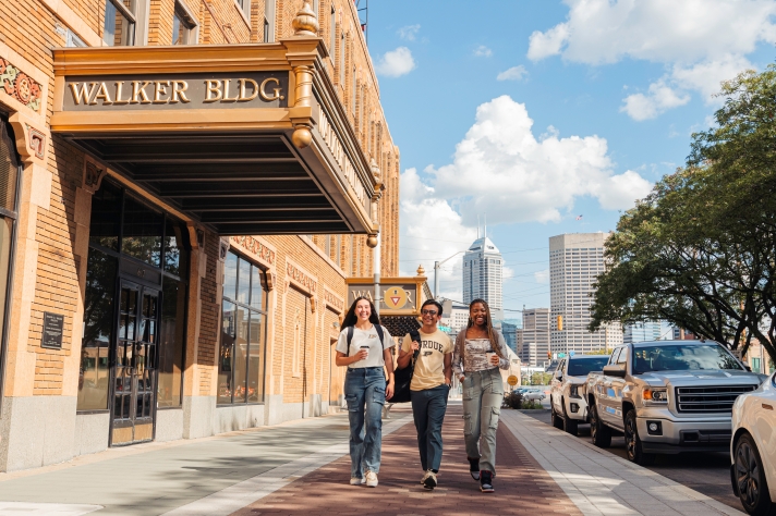 Three students walk together on a sunny sidewalk in front of Walker Building in Indianapolis, with city skyscrapers and parked cars visible in the background.