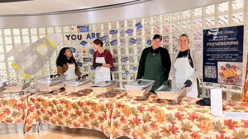 Volunteers stand behind tables covered with fall-themed tablecloths serving food for the Friendsgiving event at The Launch Pad.