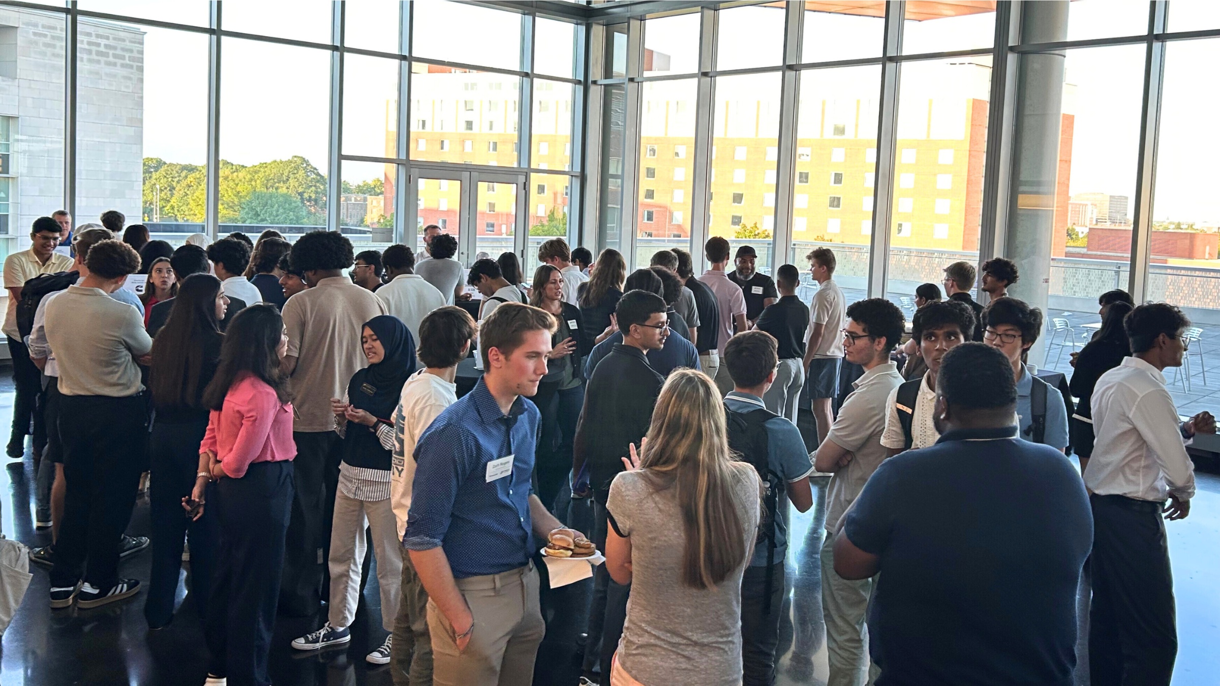 A large group of people gathered in a bright room with floor‑to‑ceiling windows, talking and standing in small groups.