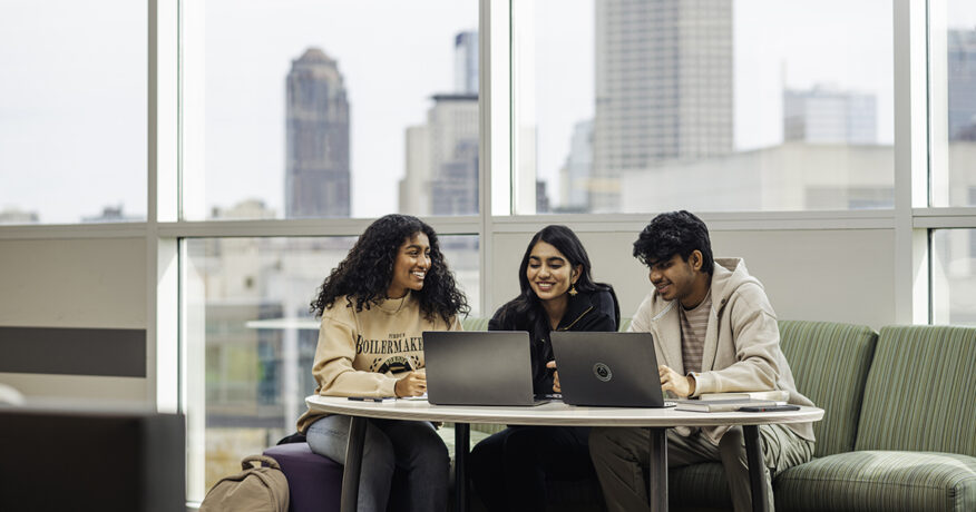 Three students sitting at a table with laptops, behind them is a window with a view of the Indianapolis city skyline