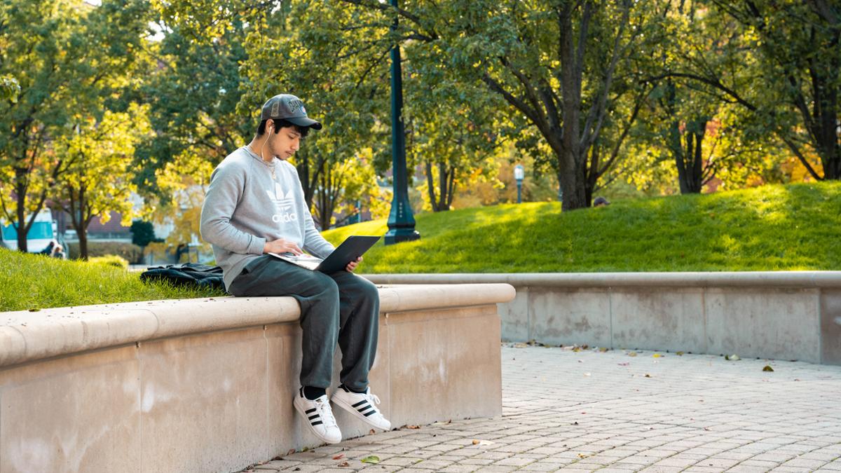 Student sitting outside on a ledge looking at a laptop computer.