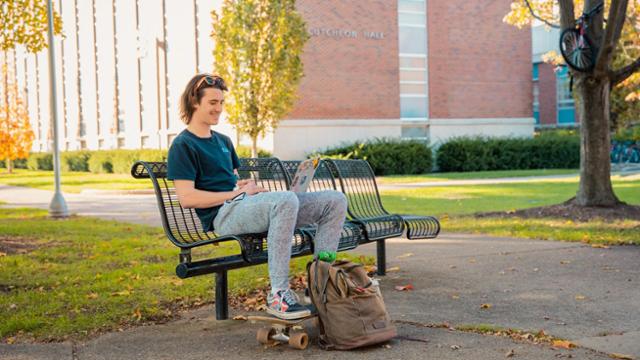 A student sitting on a bench outside working on a laptop.