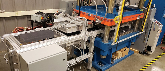 Industrial composite manufacturing setup featuring a large blue hydraulic press connected to an automated material handling and heating system, with aluminum framing, hoses, and control panels in a laboratory environment.