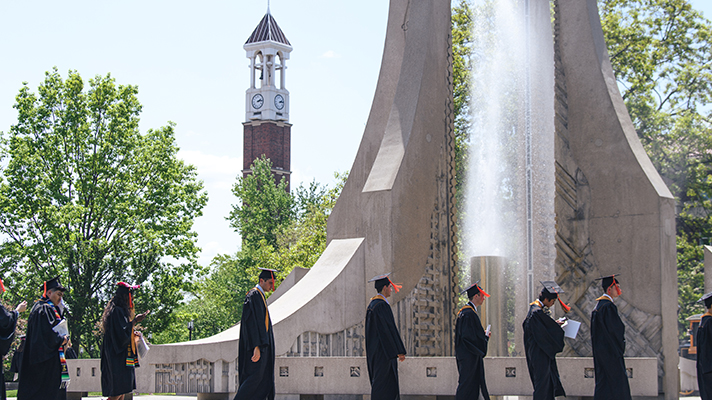 Graduates on the way to commencement ceremony walking by engineering fountain with the bell tower in the background 