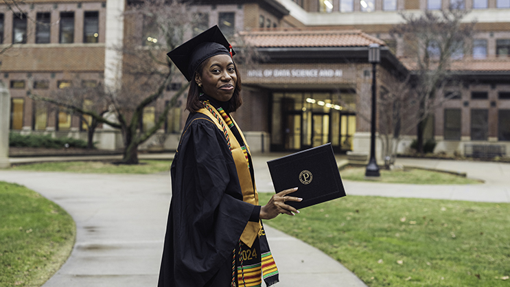 A graduate smiles holding up her diploma with the Data and AI building in the background