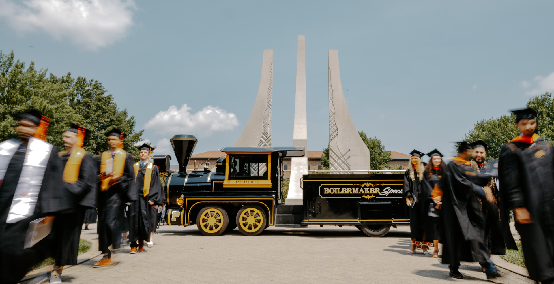 Graduations walking to commencement with the engineering fountain and the Boilermaker Special in the background