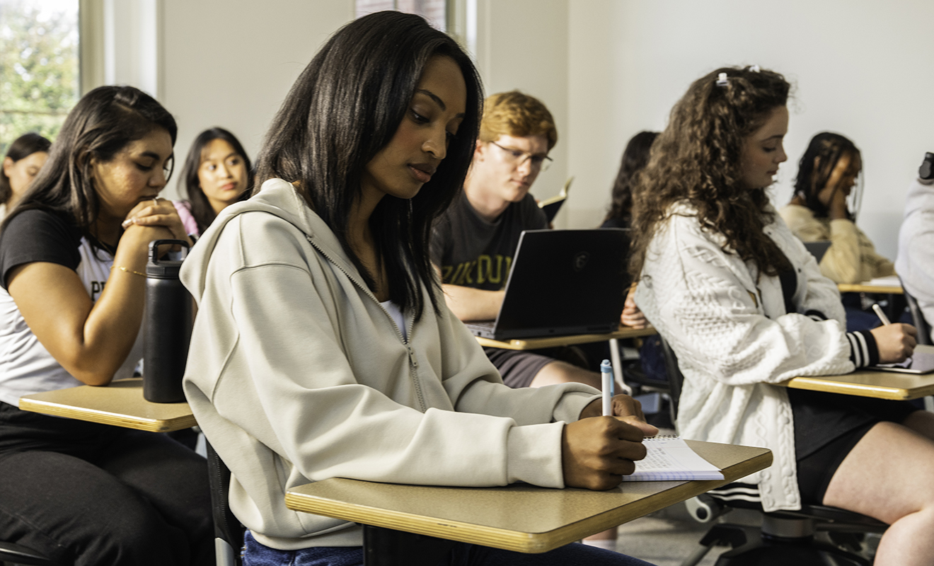 Students sit at desks in a classroom and take notes