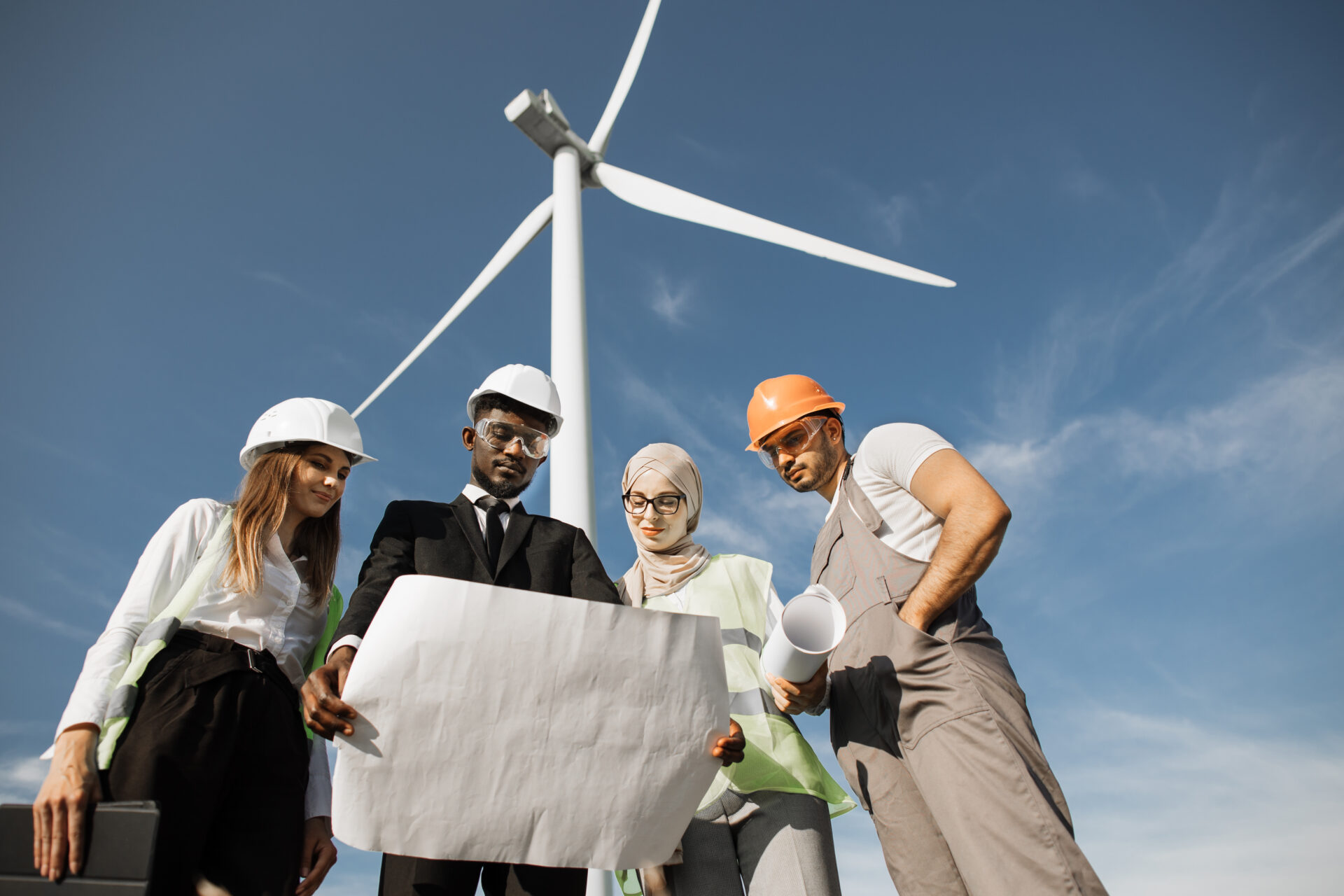 Four multi ethnic partners in safety helmets studying wind turbine blueprints while standing together at rural area. Competent industrial workers discussing common project outdoors.